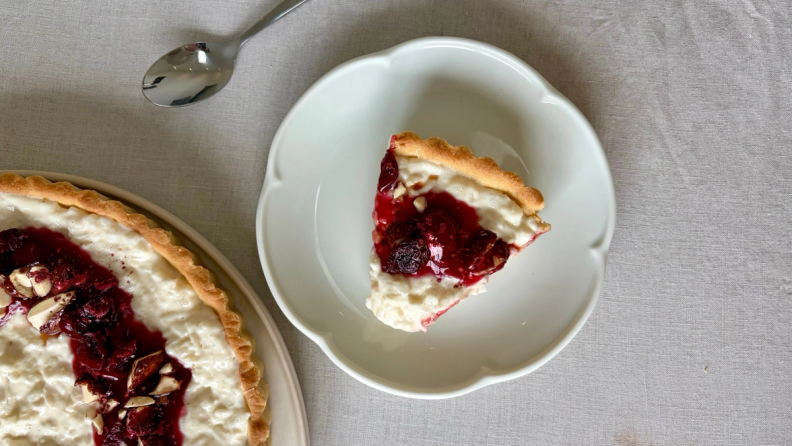 A slice of white tart with red fruit compote and almonds on a white plate, next to a larger matching tart.