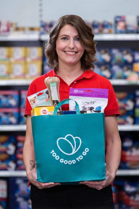 Smiling woman holding a "Too Good To Go" bag filled with pet products in a store.