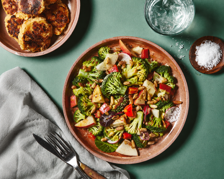 A plate of broccoli salad with apples and red peppers, a bowl of golden fritters, a glass of water, and salt on a green table.