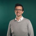 A man with a white shirt, and a gray sweater smiles against a dark green background.