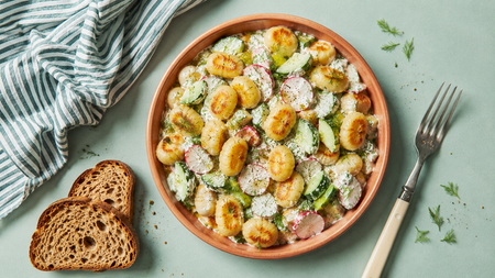 Pan-fried gnocchi with sliced radishes and cucumbers in a creamy sauce, served in a terracotta bowl with bread and a fork.