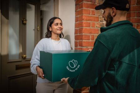 A smiling woman receives a teal "Too Good To Go" box from a delivery person.