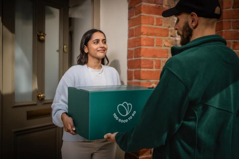 A smiling woman receives a teal "Too Good To Go" box from a delivery person.