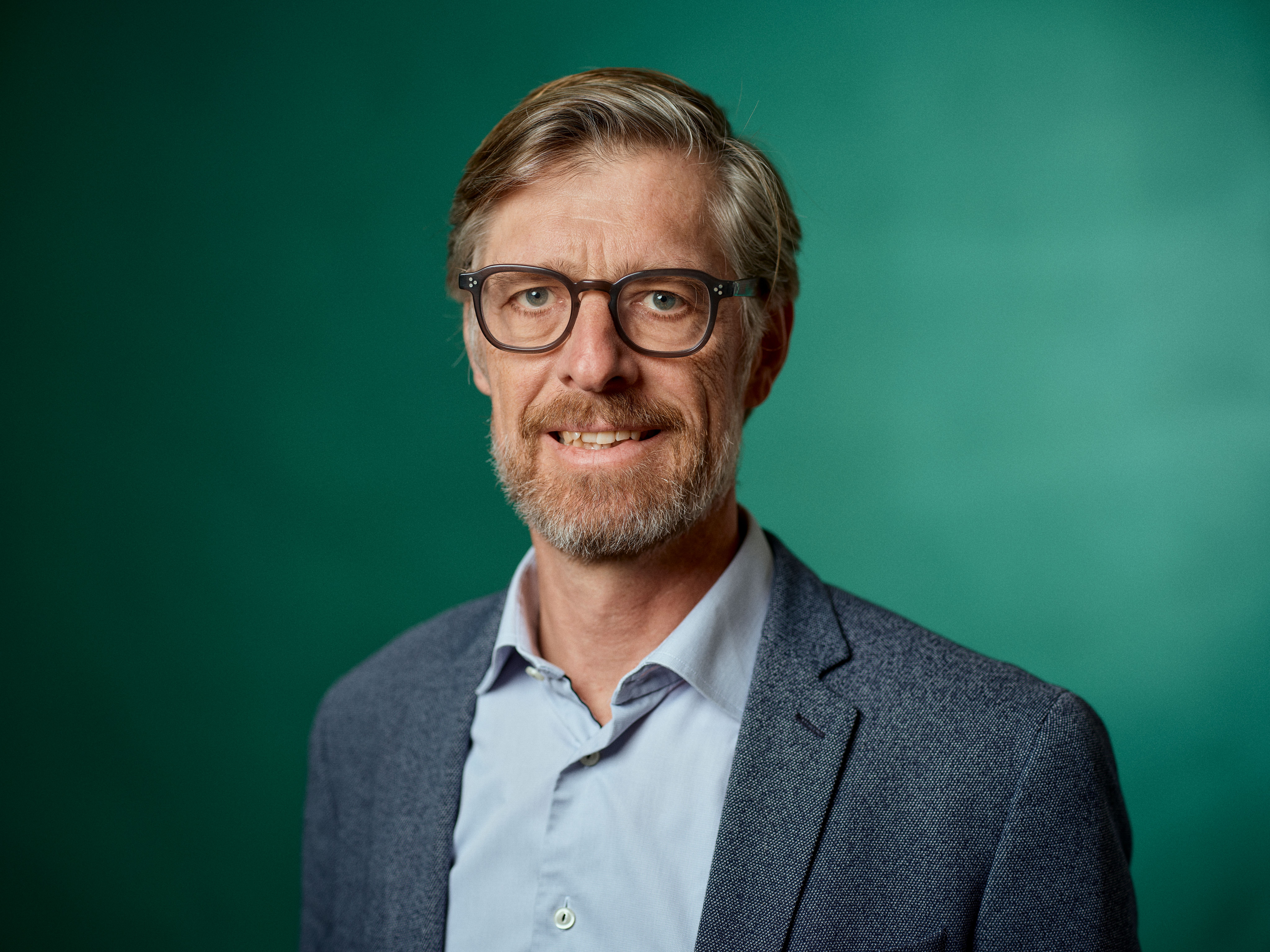 A smiling man with glasses and a beard wearing a blazer against a green background.