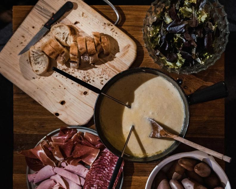 Overhead view of a cheese fondue pot surrounded by sliced bread, salad, cured meats, and small potatoes.
