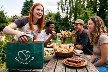 Friends share a garden picnic with food from a "Too Good To Go" bag.