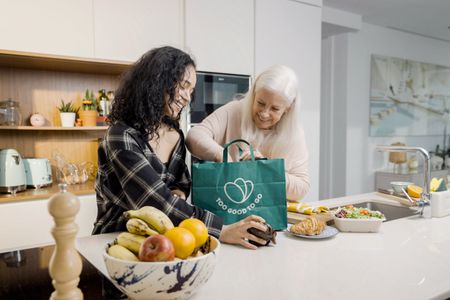 Two smiling women unpack food from a green "Too Good To Go" bag in a modern kitchen.