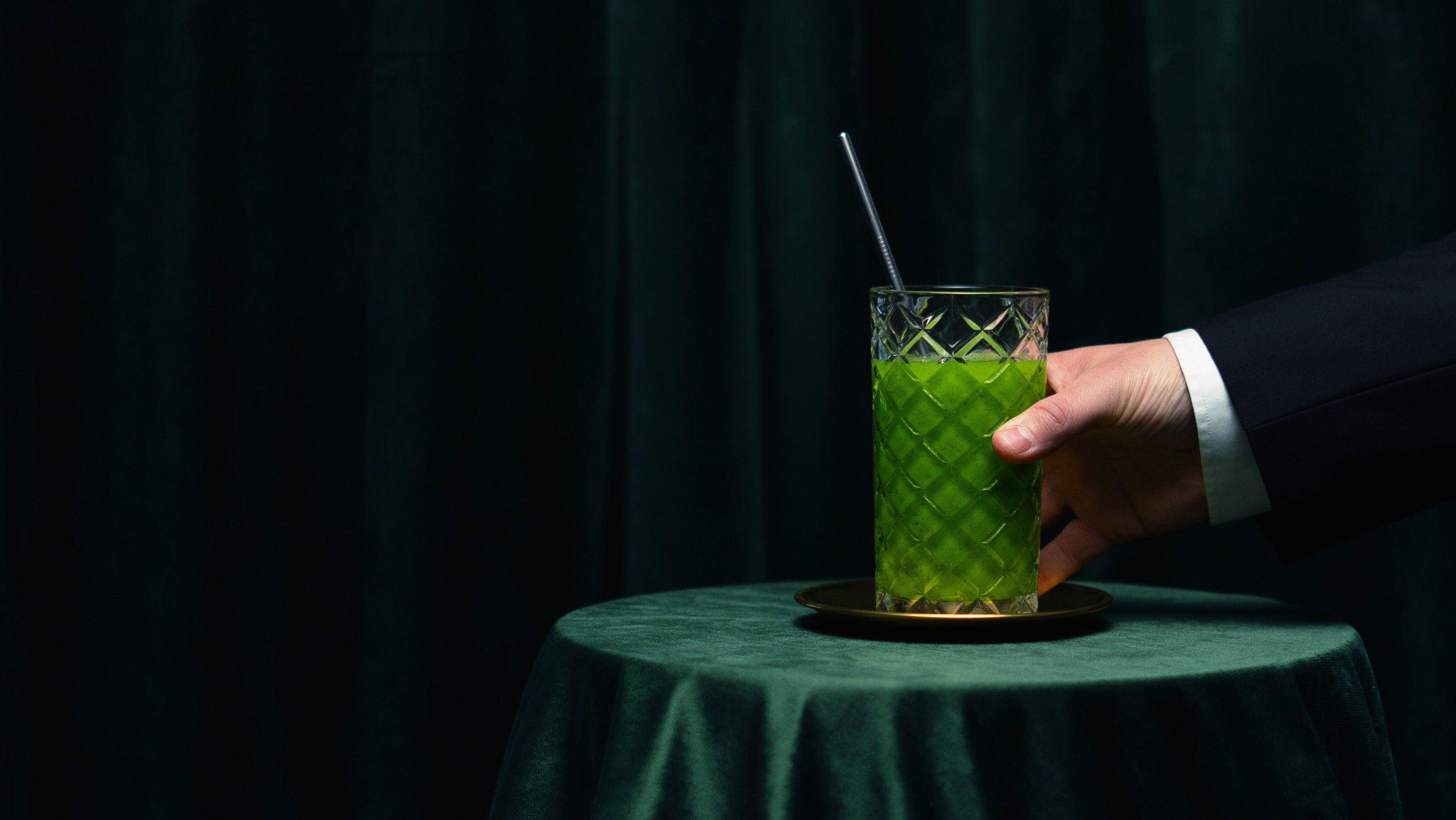 A hand in a suit jacket holds a tall glass of green liquid with a straw on a dark green velvet table.