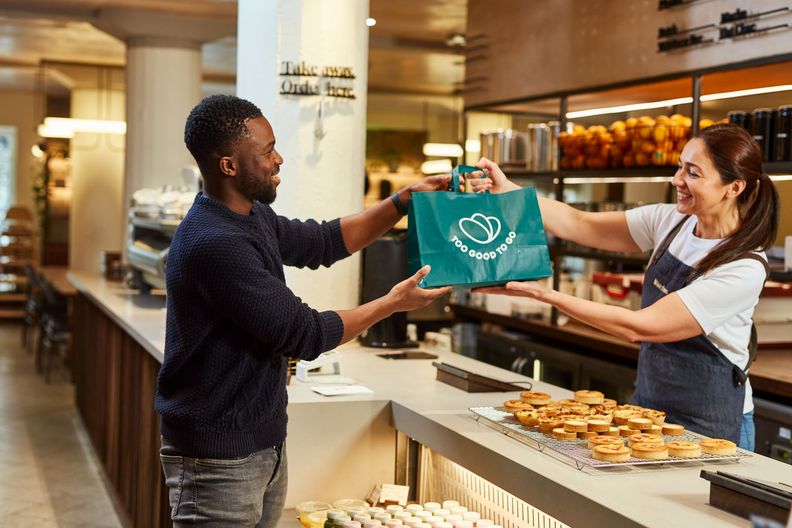 A smiling cafe worker hands a "Too Good To Go" branded bag to a smiling customer.