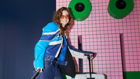 A smiling woman in a blue racing jacket and orange sunglasses pulls a white suitcase while holding a pole, against a pink tiled wall with green abstract shapes.