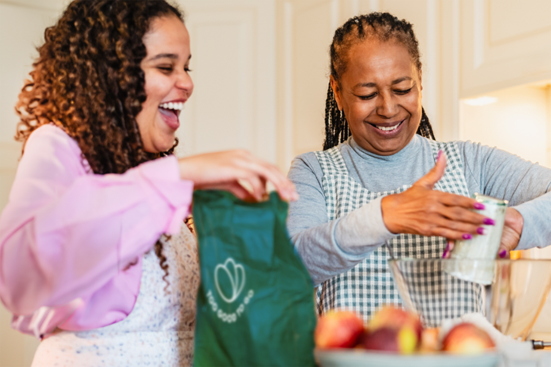Dos mujeres, vestidas con un delantal, están juntas en la cocina preparando una receta con un Pack Sorpresa de Too Good To Go
