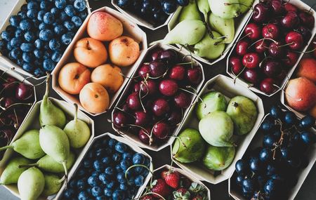 Overhead view of various fruits in small baskets, including blueberries, apricots, cherries, pears, figs, grapes, and strawberries.