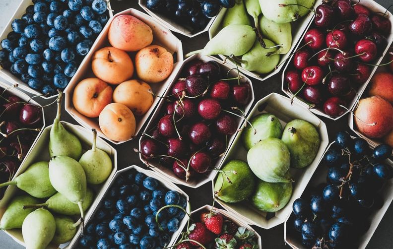 Overhead view of various fruits in small baskets, including blueberries, apricots, cherries, pears, figs, grapes, and strawberries.