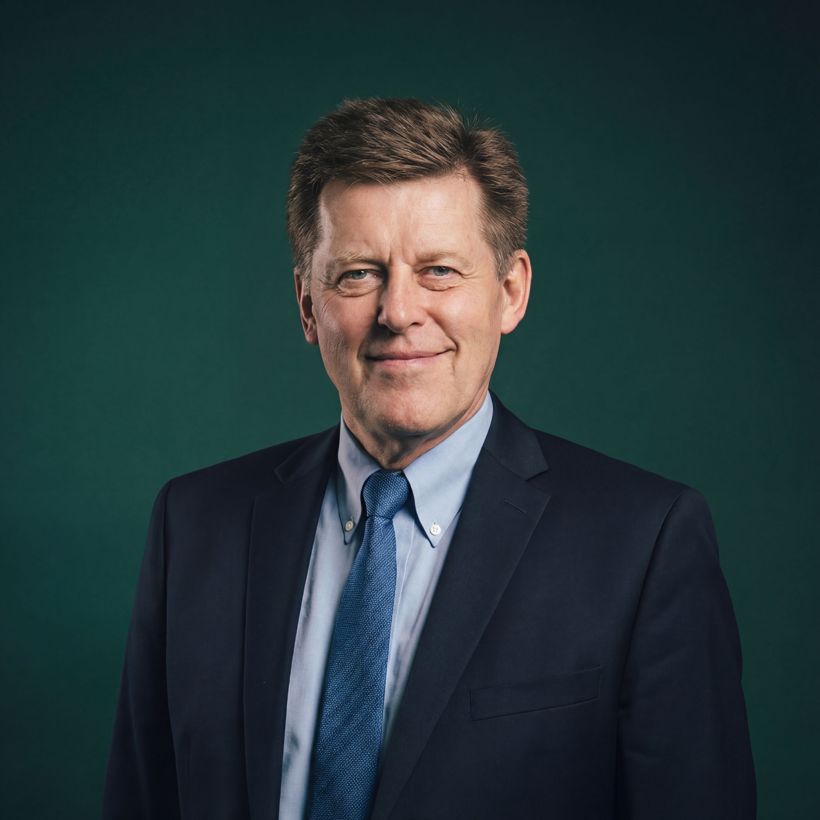 A smiling middle-aged man in a dark suit and blue tie against a dark green background.