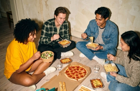 Four young adults sit on a wooden floor, smiling and eating various takeout meals including pizza, pasta, and salad.