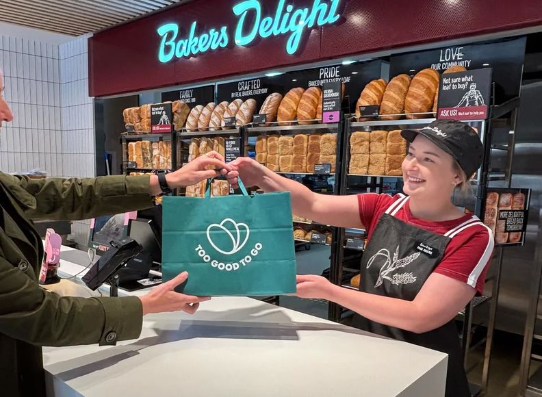 A bakery employee hands a green "Too Good To Go" bag to a smiling customer at a counter filled with bread.