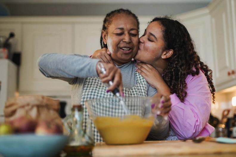 A girl kisses a woman's cheek while she bakes in a kitchen.