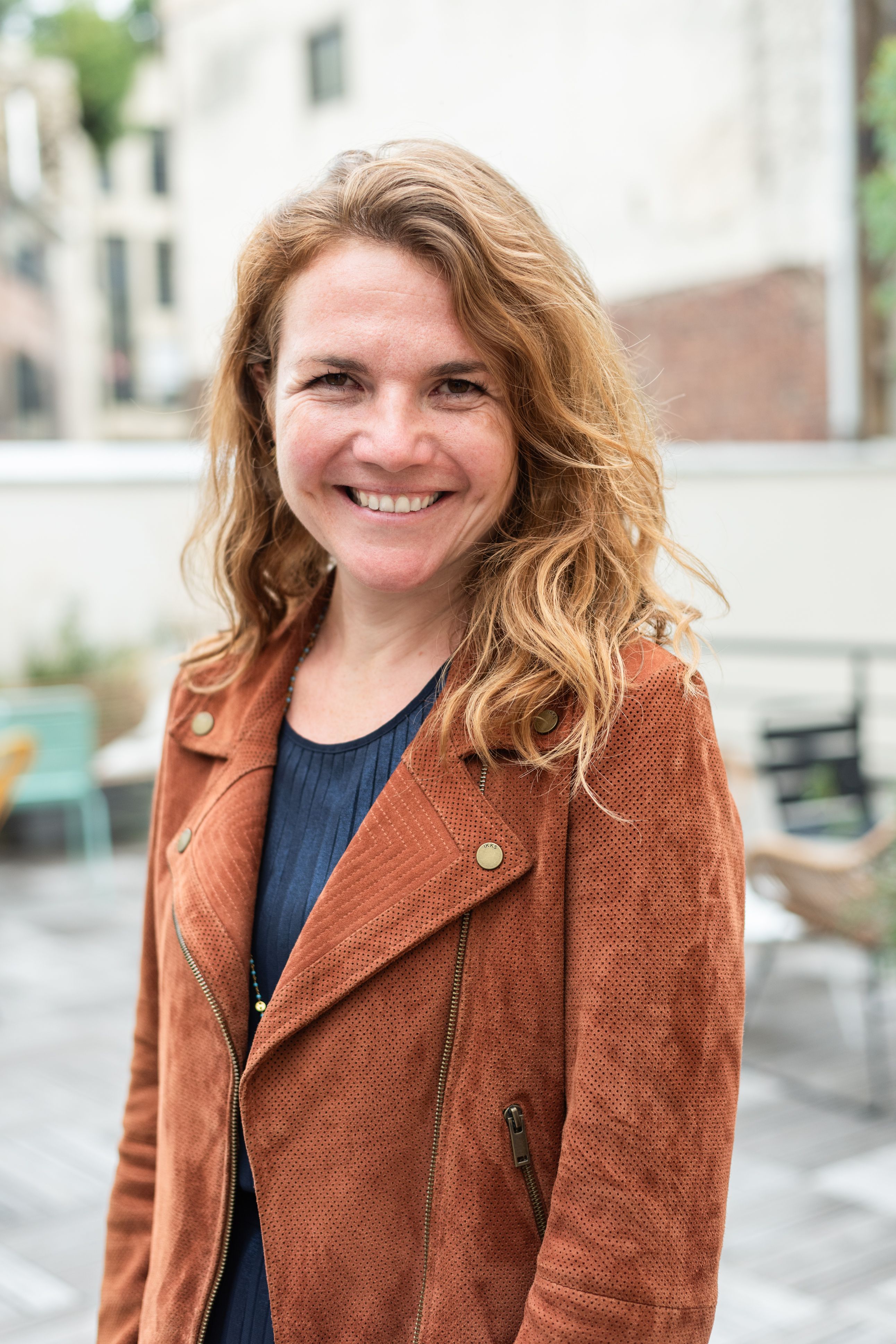 Smiling woman with wavy reddish-blonde hair wearing a brown jacket outdoors.