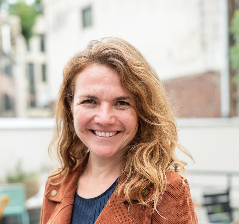 Smiling woman with wavy auburn hair wearing a brown jacket (Caroline Ray).