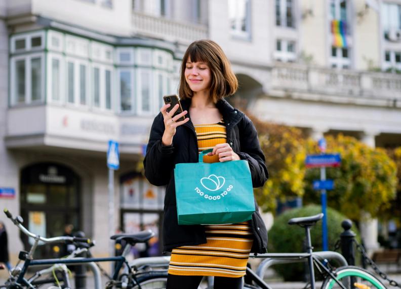A young woman smiling and looking at her phone while holding a "Too Good To Go" bag on a city street.