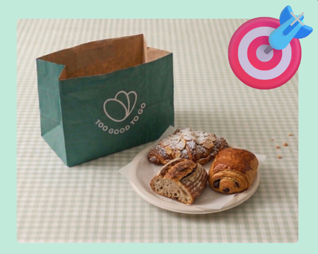 A green Too Good To Go bag next to a plate with an almond croissant, pain au chocolat, and sourdough bread, with a target and dart emoji in the corner.