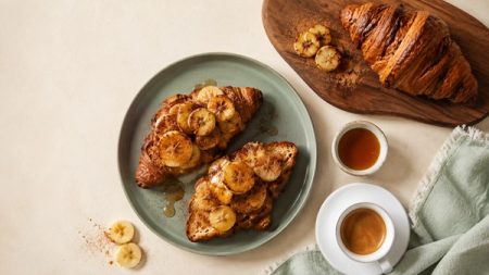 Two croissants with caramelized bananas and honey on a plate, with an espresso and another croissant nearby.