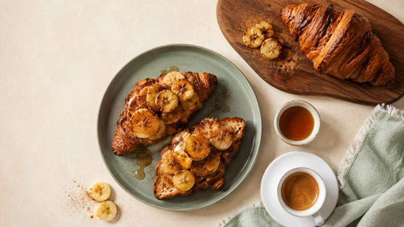 Two croissants with caramelized bananas and honey on a plate, with an espresso and another croissant nearby.