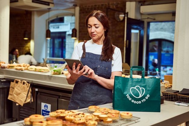 a woman is using a tablet in a bakery next to a green bag that says Too Good To Go