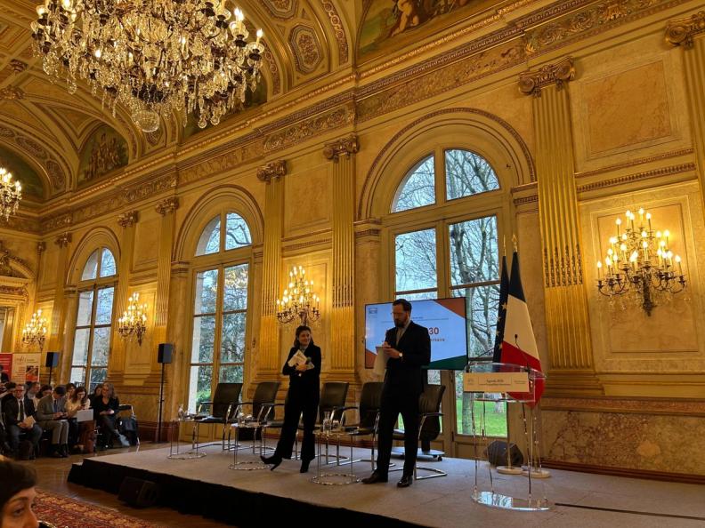 Two speakers on a stage in a grand, ornate hall with an audience, screen, and French flag.