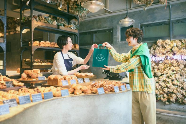 A baker hands a green shopping bag to a customer in a brightly lit bakery filled with various baked goods.