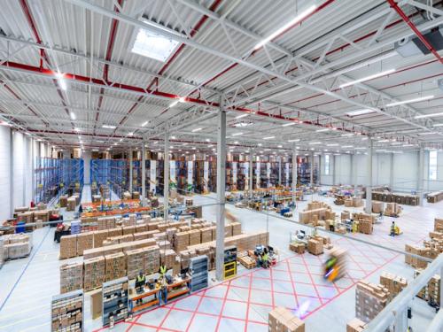 A bustling, brightly lit warehouse with rows of stacked boxes, tall shelves, and workers.