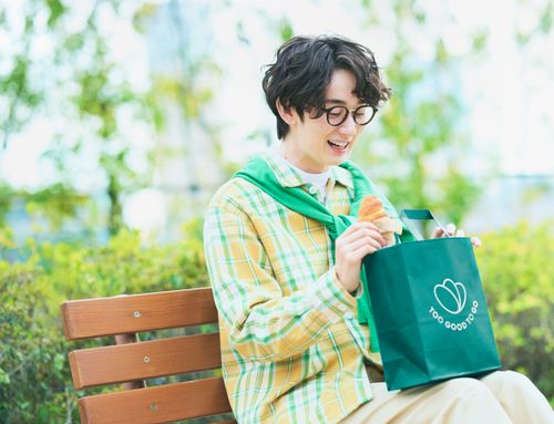 A smiling young man holds a croissant and a "Too Good To Go" bag on a park bench.