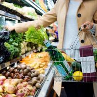 a person holding a shopping cart containing a lemon, a water bottle and other grocery items, picking up greens from the supermarket shelves