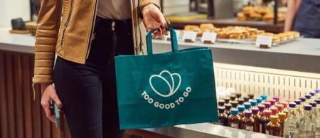 A person holds a green "Too Good To Go" branded bag in a cafe.