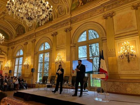 Two speakers on a stage in a grand, ornate hall with an audience, screen, and French flag.