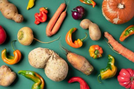 A flat lay of various misshapen vegetables, including carrots, potatoes, peppers, celeriac, and a pumpkin, on a green background.