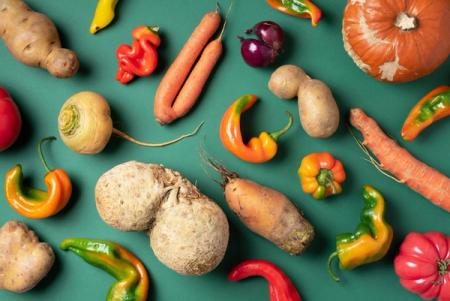 A flat lay of various misshapen vegetables, including carrots, potatoes, peppers, celeriac, and a pumpkin, on a green background.