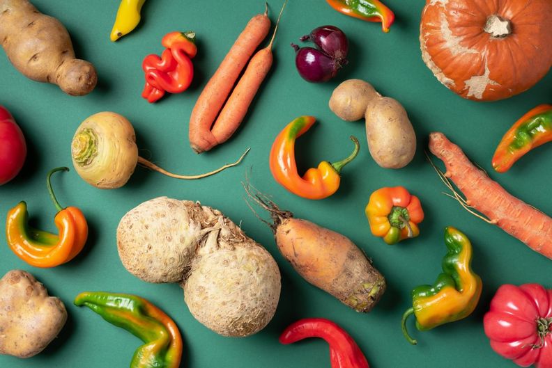 A flat lay of various misshapen vegetables, including carrots, potatoes, peppers, celeriac, and a pumpkin, on a green background.