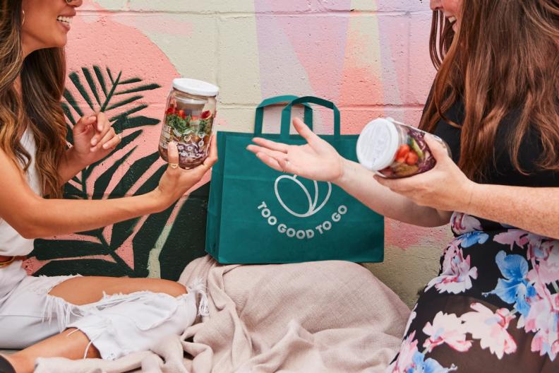 two women are sitting on a blanket holding jars of food .