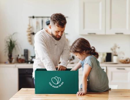 A man and a young girl looking into a green "Too Good To Go" box in a kitchen.