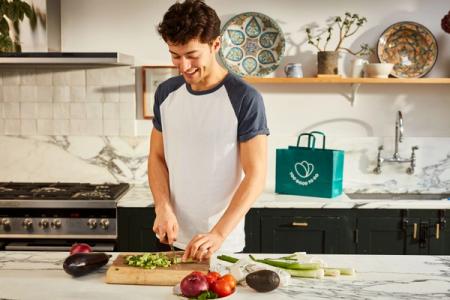 A happy young man chops vegetables in a kitchen next to a green "Too Good To Go" bag.