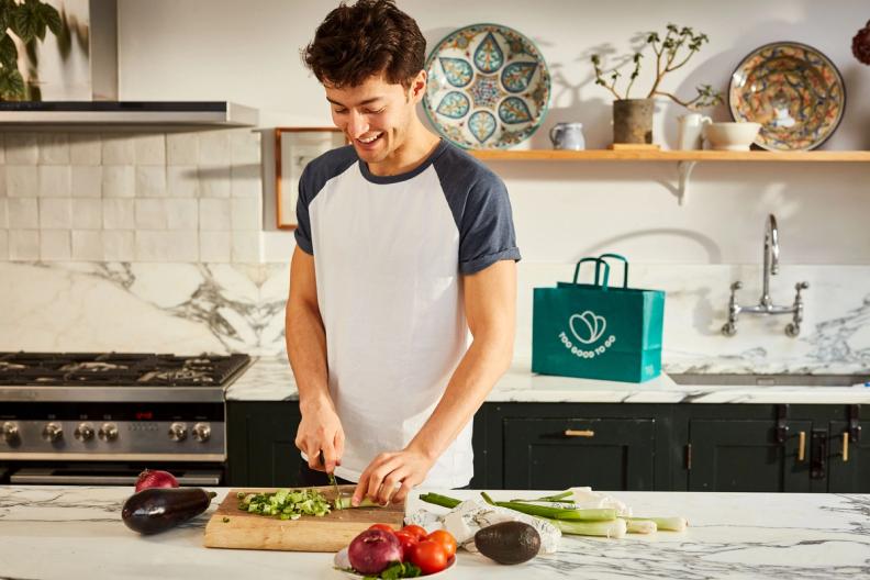A happy young man chops vegetables in a kitchen next to a green "Too Good To Go" bag.
