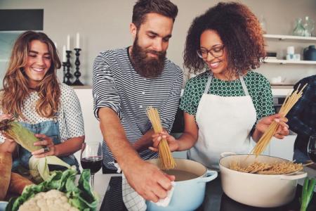 Three smiling people cooking together in a kitchen, preparing pasta and vegetables.