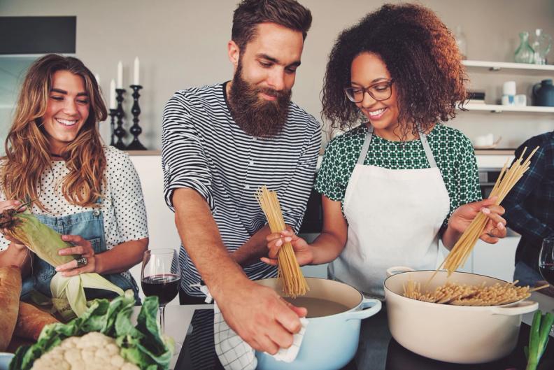 Three smiling people cooking together in a kitchen, preparing pasta and vegetables.