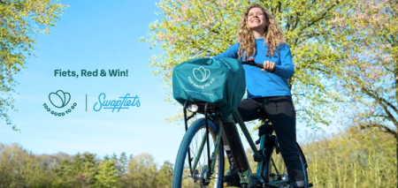 A smiling woman on a bicycle with a Too Good To Go front basket cover, in a sunny park. Logos for Too Good To Go and Swapfiets are visible, alongside the text "Fiets, Red & Win!".