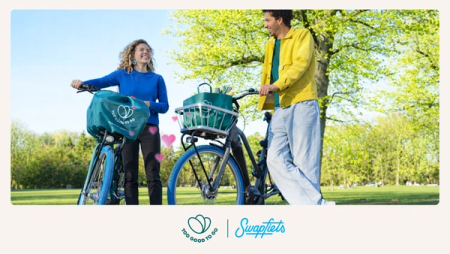 A smiling man and woman with Too Good To Go branded Swapfiets bicycles and pink hearts in a sunny park, with Too Good To Go and Swapfiets logos at the bottom.