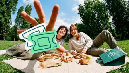 Two happy women picnic in a sunny park with food. An overlaid hand presents two green tickets.