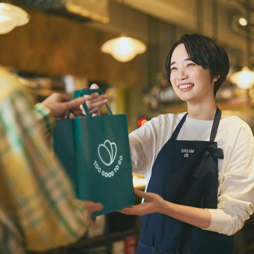 A smiling woman hands a "Too Good To Go" bag to a customer.
