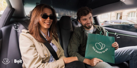 A smiling man and woman in a car, with the man holding a green "Too Good To Go" bag.