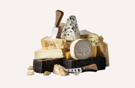 A selection of cheeses including blue cheese, brie, and hard cheeses, with two cheese knives on a dark cutting board.